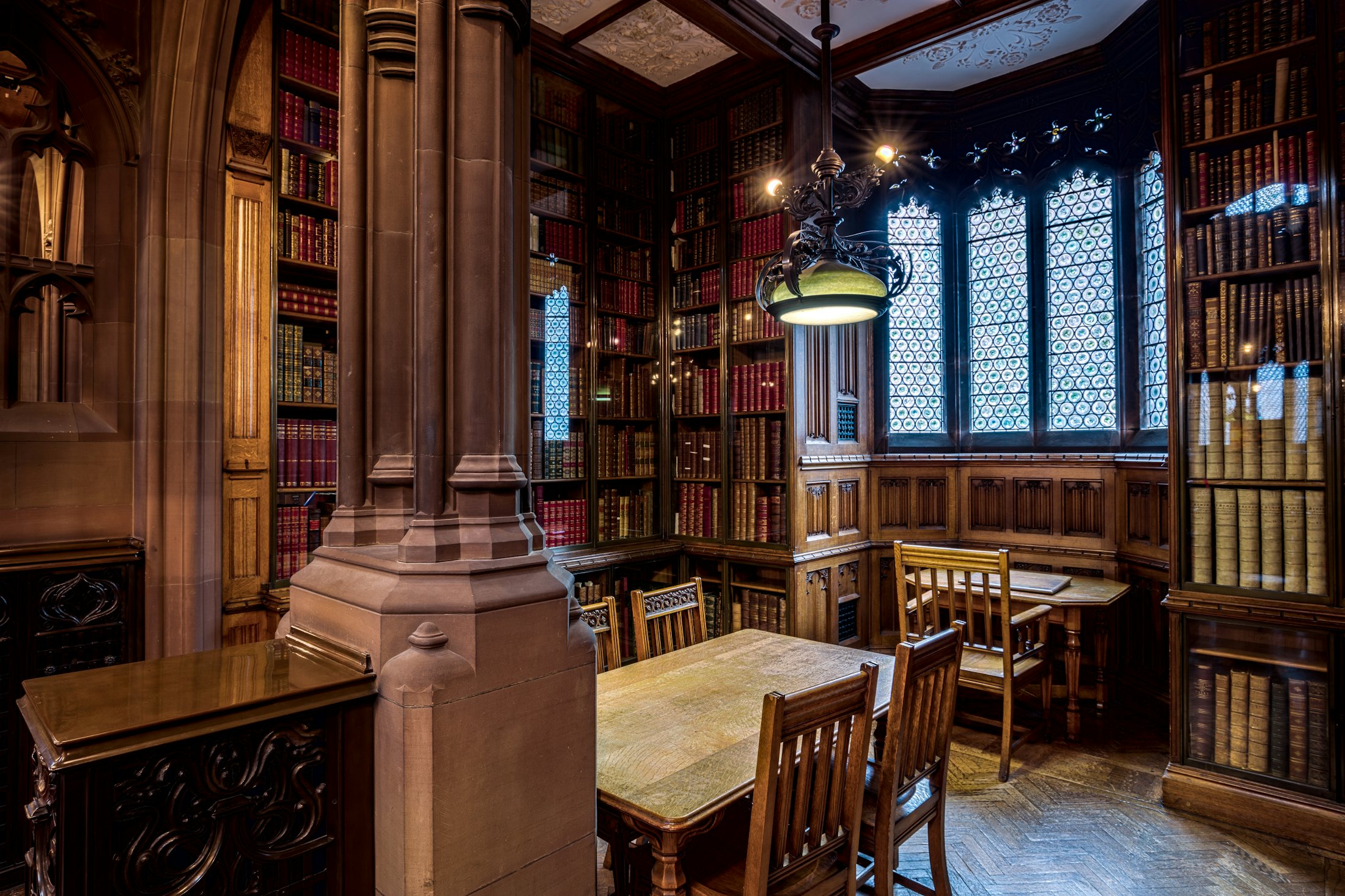 A wood-panelled library reading room with carved bookshelves, stone pillars, leaded windows, and study tables.